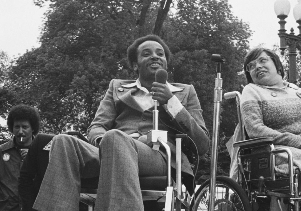 Brad Lomax addresses a crowed while holding a microphone with a smile on his face.  The photo from the 1970s in in black and white. Mr. Lomax, and African-American man who uses a wheelchair is wearing a plaid bellbottom suit with wide lapels. 