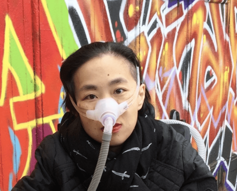 Alice Wong gazes pensively at the camera. She is wearing a black coat and scarp with white graphic designs.  She is photographed in front of bright red and orange graffitied wall.