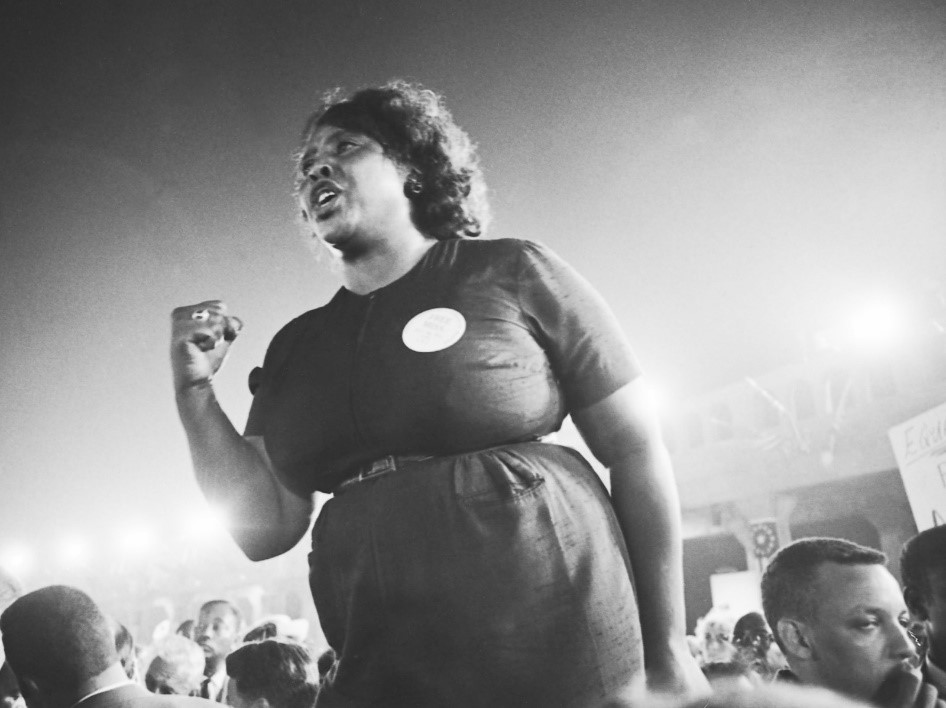 A black and white image of Fannie Lou Hamer at a rally or protest. She is backlit by bright lights, and has one fist raised to shoulder height, as she speaks.