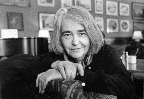 A black and white photo of Kate Millett with her hands folded stares confidently into the camera. 