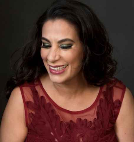 A photographic portrait of Maysoon Zayid. She has shoulder length brown hair, is smiling and wearing a maroon garment.