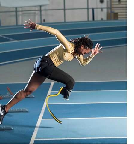 Femita begins a race, wearing a yellow shirt and black running shorts. Her arms are in a running motion, and she has a prosthesis on her right leg. 