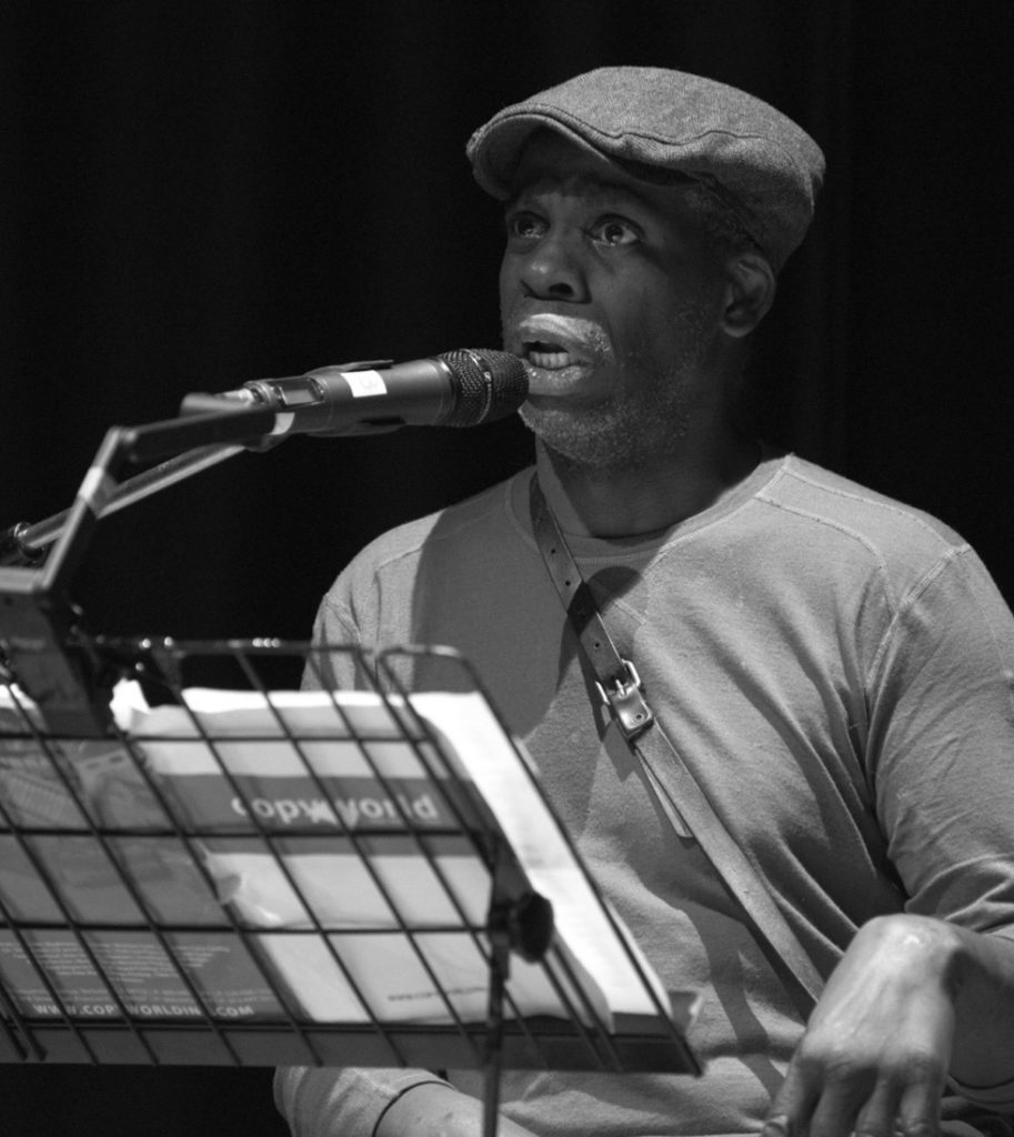 A black and white photograph of Leroy F. Moore Jr., an African American man with a short silver flecked goatee, performing spoken-word poetry. Mr. Moore speaks into a microphone with a music stand in front of him. He is wearing a flat cap, a long sleeve tee, and a cross body bag with a leather strap. 