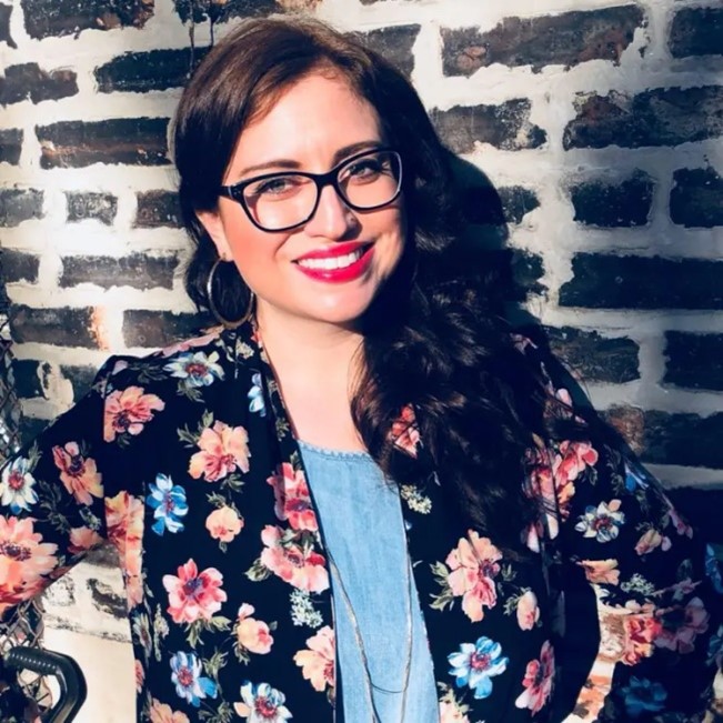 Katherine Perez, Co-Founder, The National Coalition for Latinx with Disabilities, stands against a brick wall, smiling. She has brown hair, glasses, and is wearing a flowered jacket.