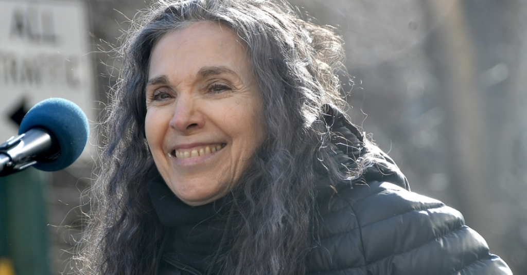 Nadina LaSpina speaks at a microphone. She is smiling, has curly gray hair, and is wearing a black winter coat.