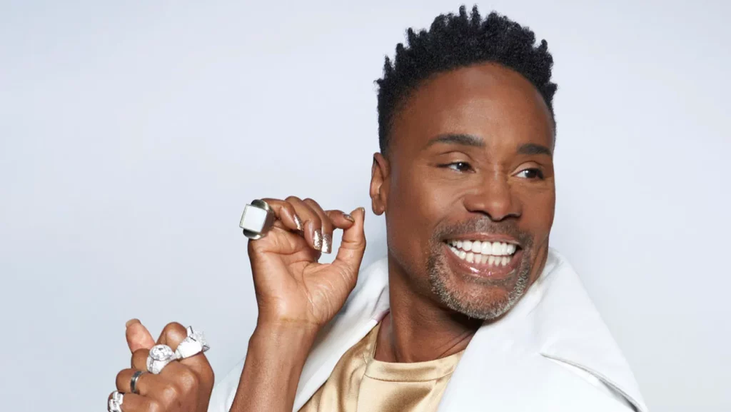 A photo from the shoulders up, Billy Porter stands smiling in front of a white background. He is an African American man, with short black hair. He is wearing a white jacket and has rings on several fingers. 