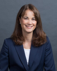 A headshot of Rachel Kaprielian, who has shoulder length dark brown hair, and is wearing a dark blue jacket over a white shirt. She is smiling.