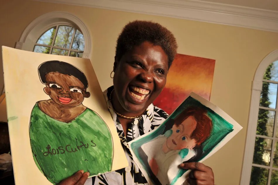 Lois Curtis stands in a room holding two paintings she painted. She is a Black woman with short black hair, wearing a black and white shirt. One painting is a self-portrait and the other is of another woman. She has a big smile.