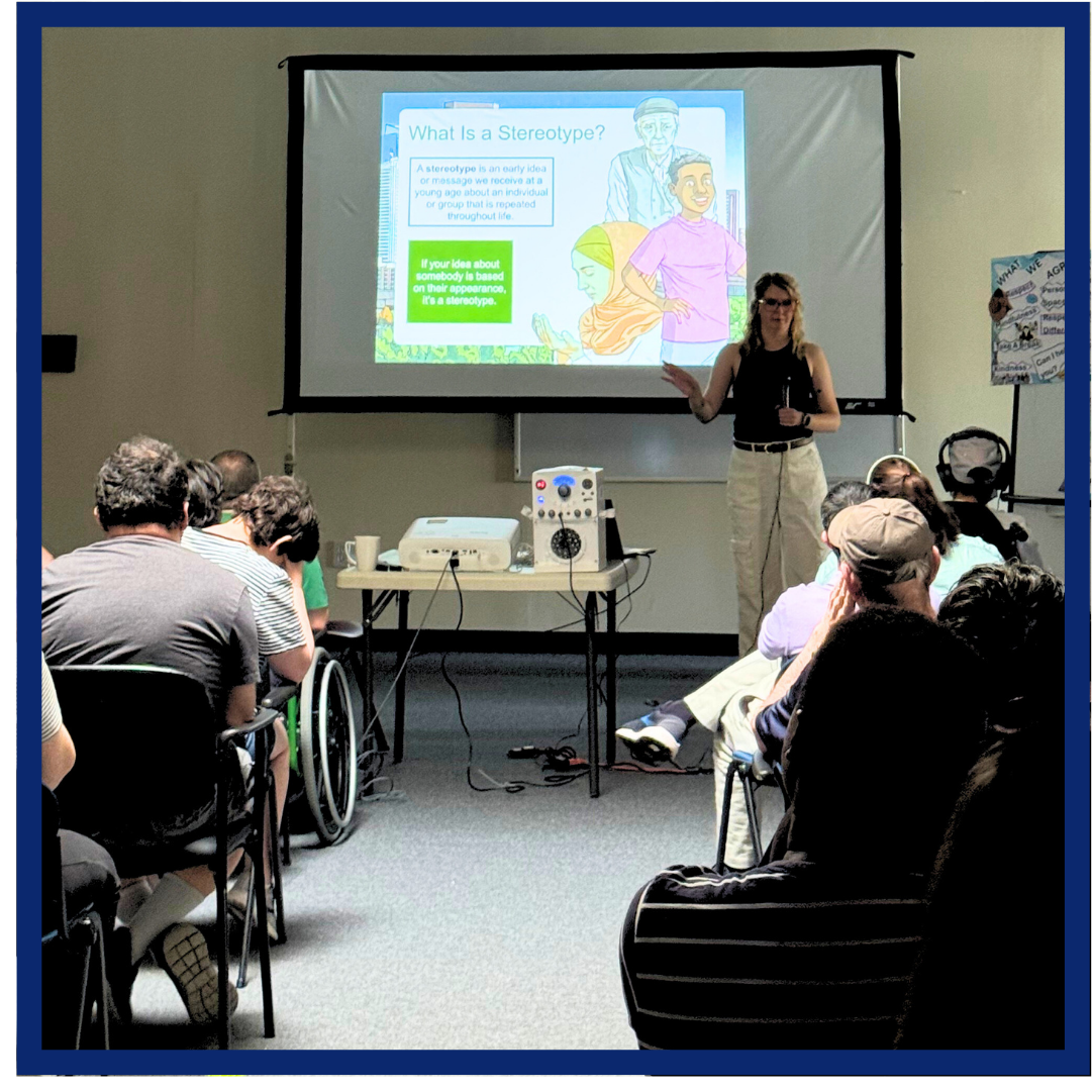 Melissa (in a black tank top and white pants) stands in front of a room of students, teaching with a powerpoint presentation behind her.
