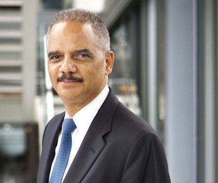 Eric Holder stands facing the camera, wearing a black suit and blue tie. He is a Black man with short gray hair and a black mustache.