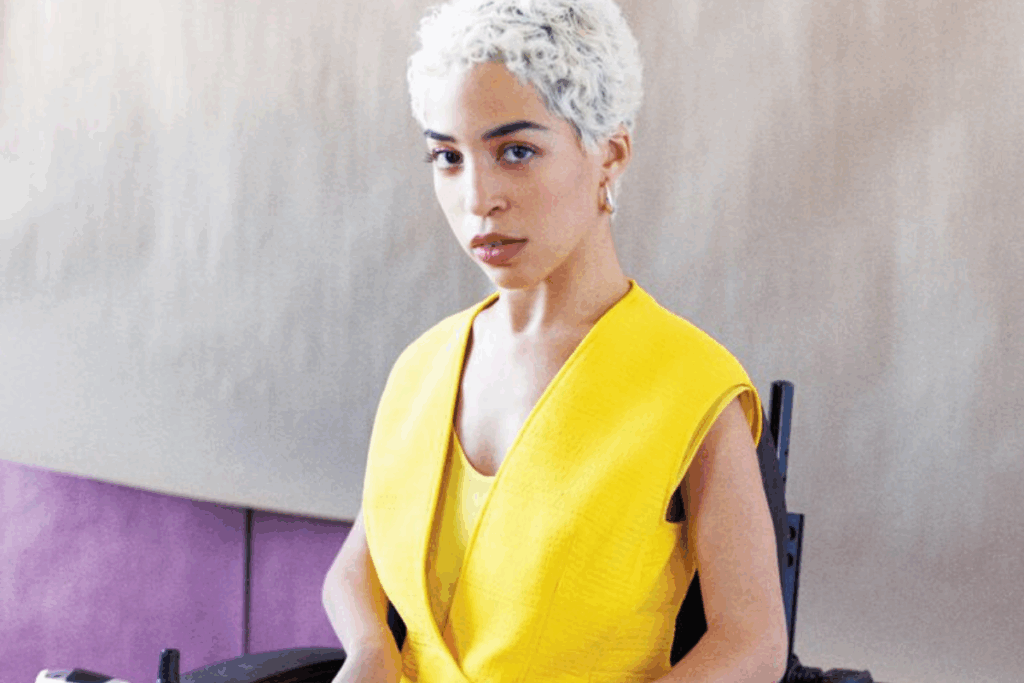 Jillian Mercado, a young woman with short white hair and dark eyes, sits in a bright yellow dress in a wheelchair.