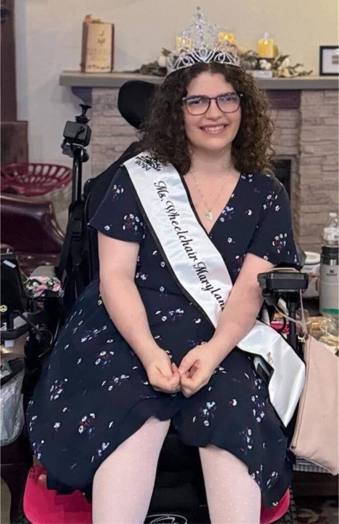 Dominique Sessa sits in a wheelchair, wearing a black dress with white flowers, a tiara, a sash saying "Ms. Wheelchair Maryland," and she has dark shoulder-length hair and glasses. She is smiling.