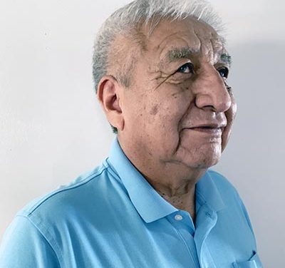 Hoskie Benally Jr. stands with a slight smile at an angle in front of a white background. He is wearing a blue polo shirt, and has short white hair.