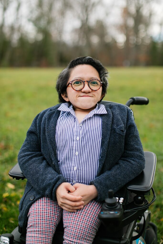 Sandy Ho sits in a wheelchair with her hands clasped in front of her. She has short brown hair, wears glasses, and a striped shirt with a sweater over it.
